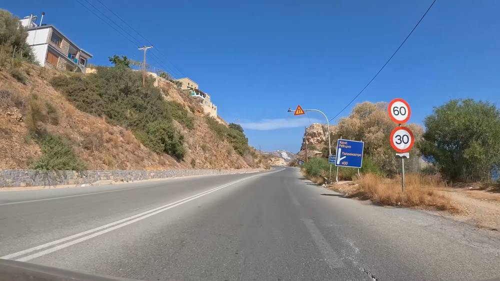 A scenic road in Crete, Greece, showing speed limit signs of 60 km/h and 30 km/h, along with a directional road sign. The image highlights the local traffic regulations and road signage in a mountainous area under a clear blue sky.