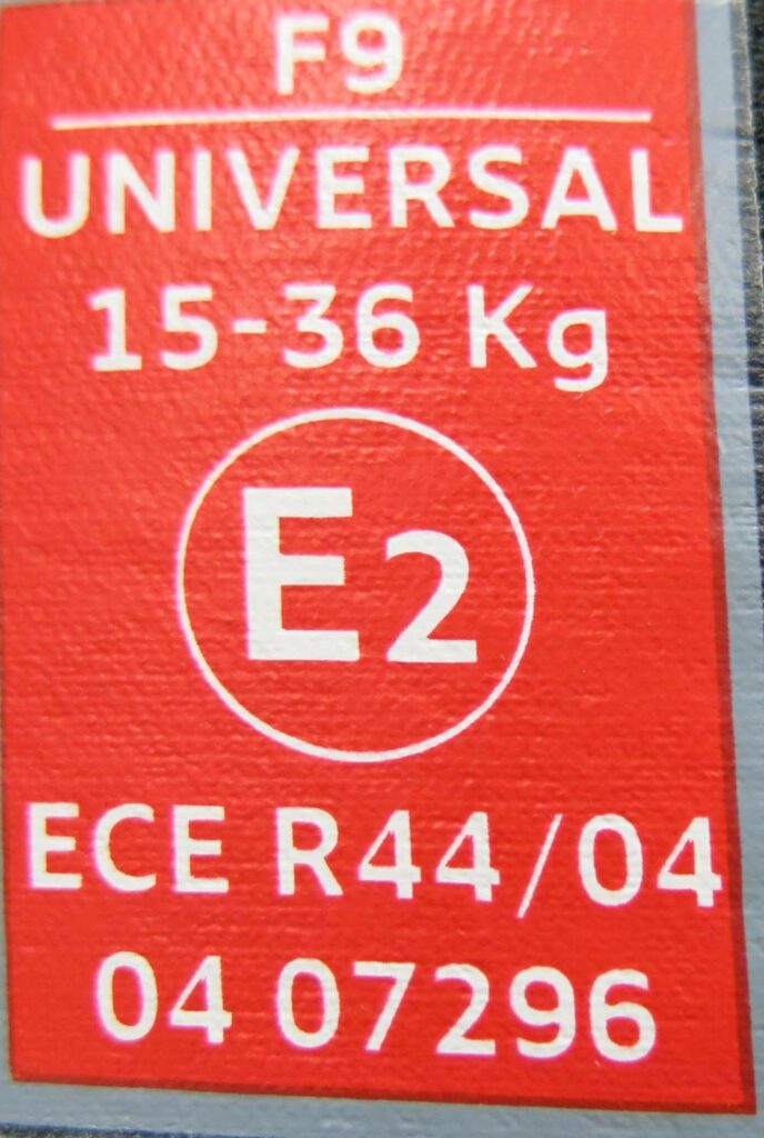 A red ECE R44/04 certification label for a child car seat, indicating universal compatibility for children weighing 15-36 kg, with the code E2.