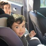 Two children sitting in the back seat of a car, one in a forward-facing car seat and the other in a booster seat, ensuring safe travel