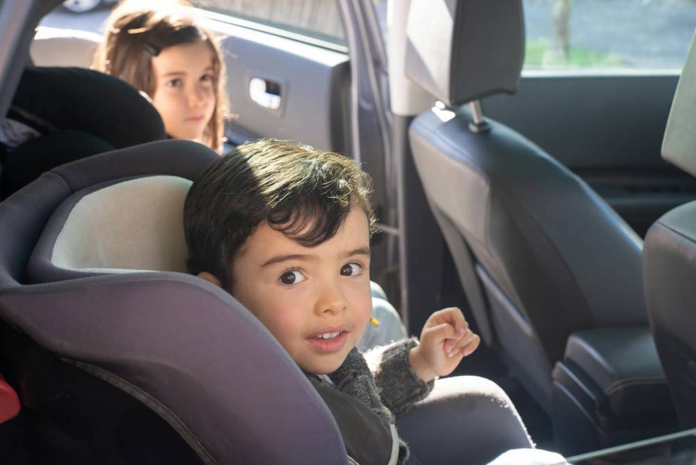 Two children sitting in the back seat of a car, one in a forward-facing car seat and the other in a booster seat, ensuring safe travel